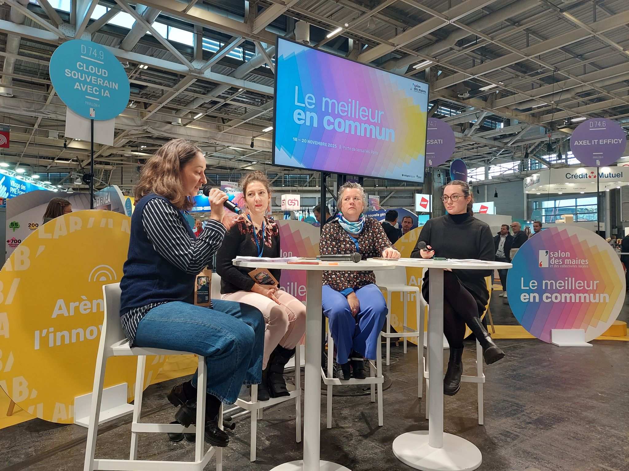 photo de quatre femmes assises autour d'une table blanche, dont une tout a gauche qui parle micro à la main, avec écran projetant le slogan "le meilleur en commun"